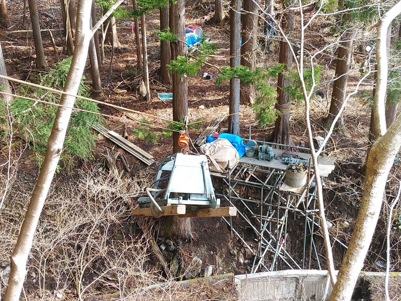 山林の斜面に足場を組み、ワイヤーで機材を吊り下げてボーリング調査を行っている様子