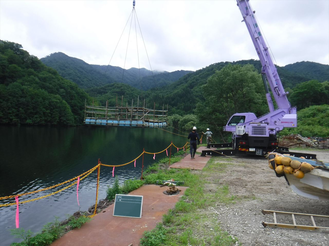 ダム湖上でクレーン車を使い、水上に足場やぐらを設置している様子。作業員2名がロープを持ちながら岸沿いを移動している