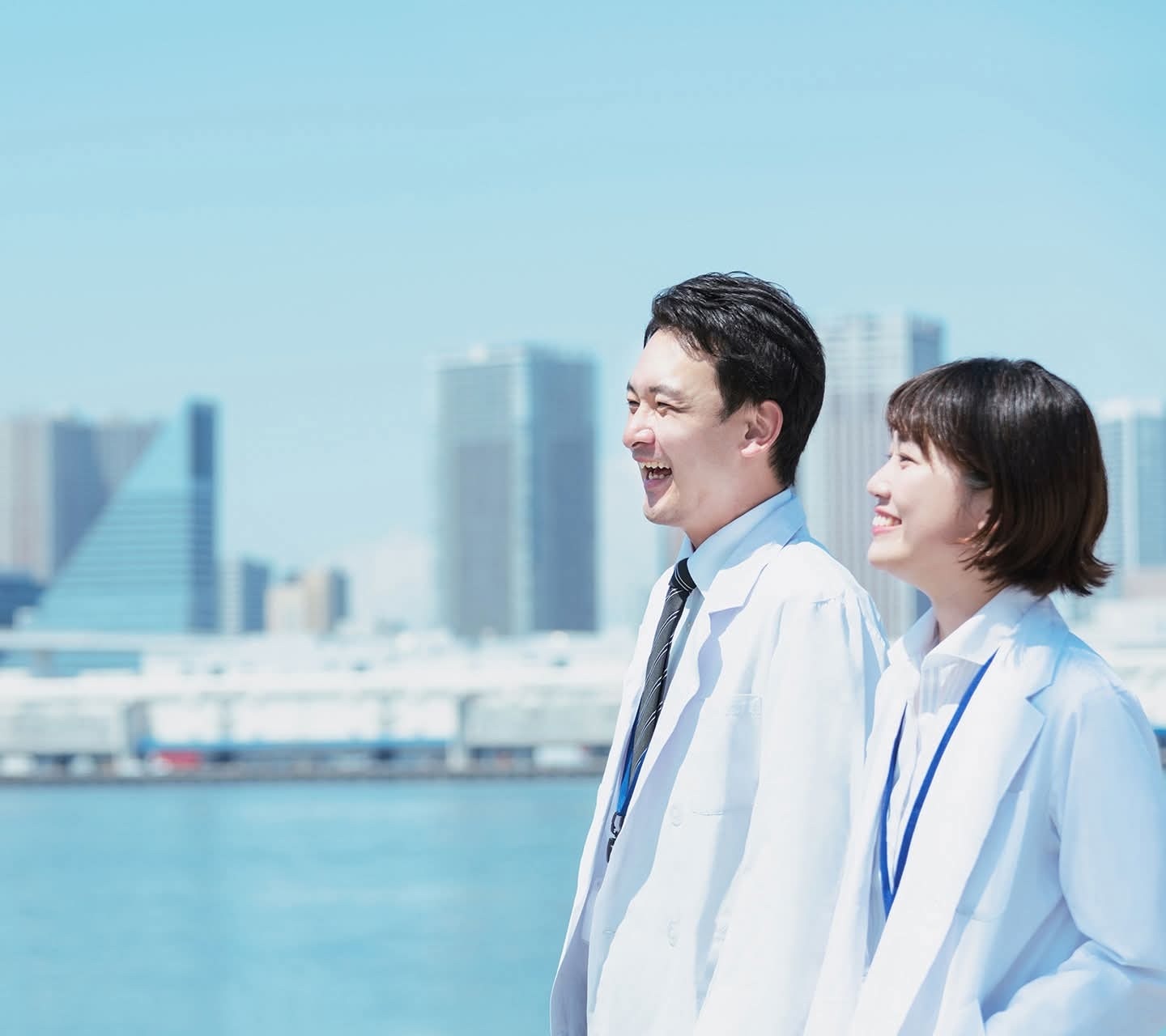 Two professionals in white coats smile while walking along a waterfront with a city skyline in the background