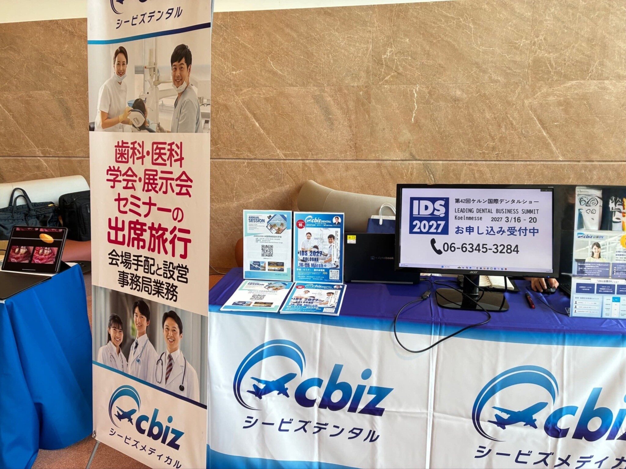 「シービズデンタル」の展示会ブース。歯科・医科学会・展示会・セミナーの出席旅行、会場手配と設営、事務局業務と書かれたのぼりと、案内が表示されたモニター、チラシが並ぶ机が設置されている