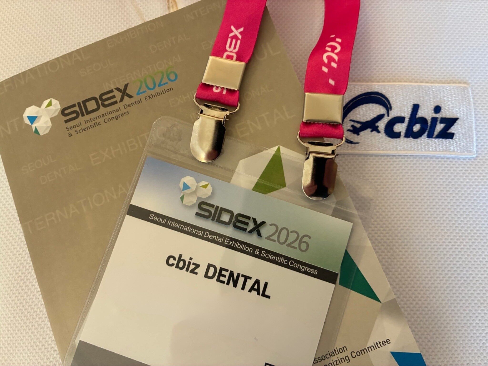 「cbiz DENTAL」と記載されたSIDEX 2026の来場者証と、cbizのネームタグ付きピンク色のストラップ、SIDEX 2026のパンフレットが並んでいる