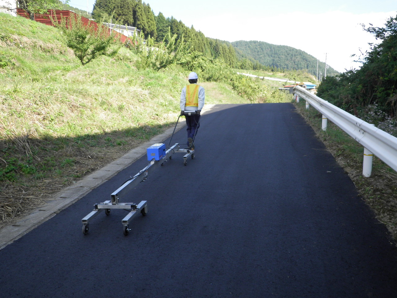 舗装された道路上で、作業員が牽引式の計測機器を引きながら移動している様子