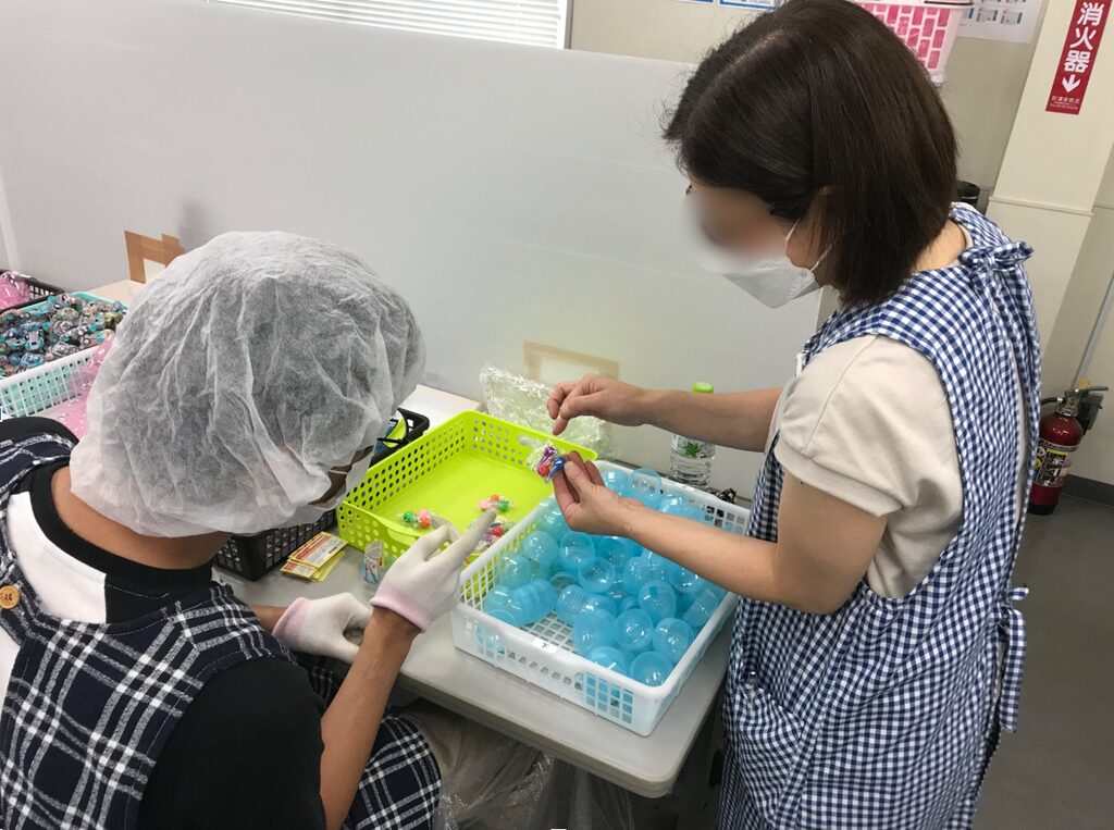作業台の上で、2人の人物がカプセル容器や小袋を扱っている。片方はヘアキャップと手袋を着用し、もう片方はマスクとエプロンを着用している。作業台には青いカプセル容器が入ったかごや、小物が入ったかごが置かれている。
