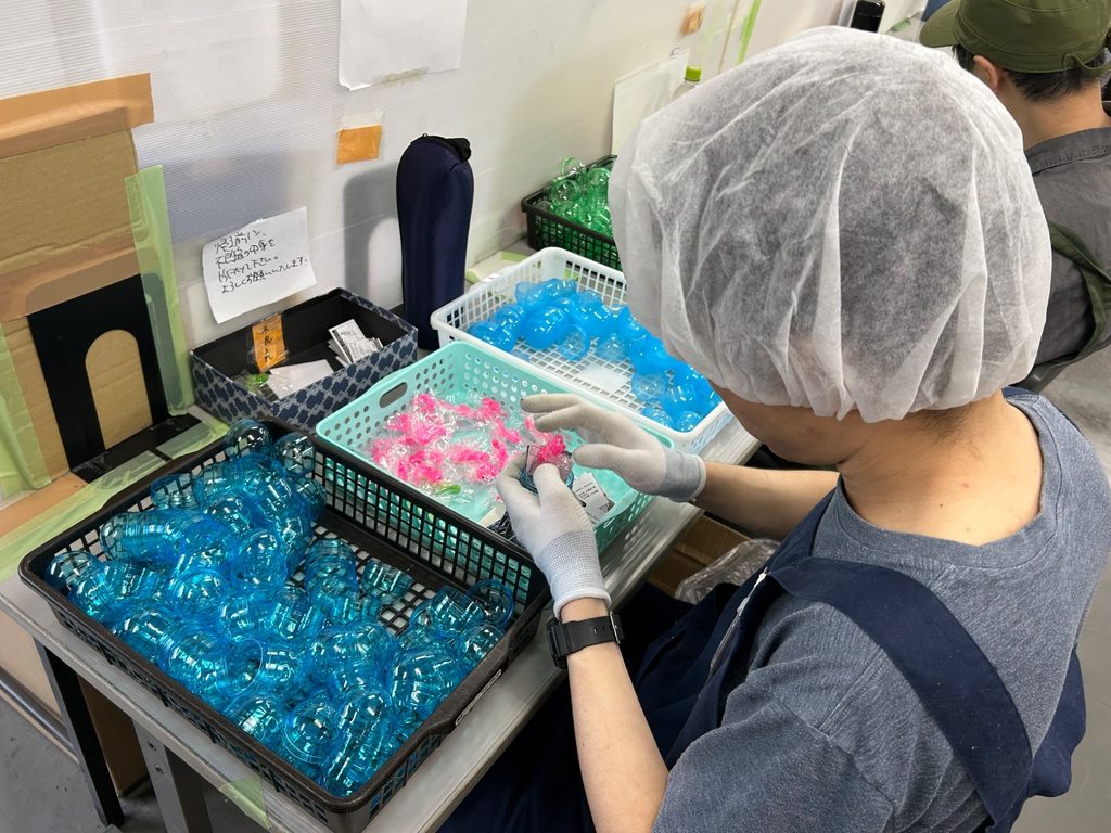 ヘアキャップと手袋を着用した人物が、作業台の上で小袋を扱っている。作業台には青いカプセル容器が入ったかごや、ピンク色の小物が入ったかごが置かれている。奥には別の人物が座っている。