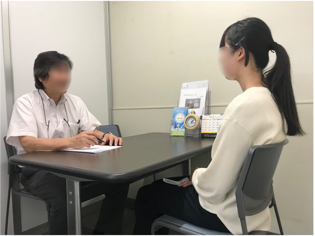 机を挟んで、白いシャツを着た男性と白いセーターを着た女性が向かい合って座っている。男性は紙の上に手を置き、女性は椅子に座って男性の方を向いている。机の上には書類、卓上カレンダー、時計のような物が置かれている。