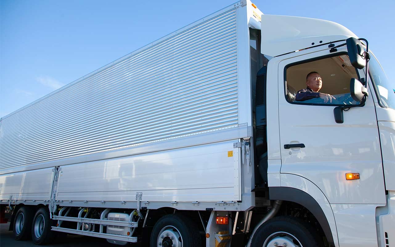 白い大型トラックの運転席に人が座っており、青空の下で撮影されている。