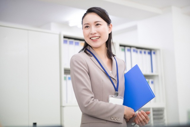 オフィス内で、青いファイルを持ったスーツ姿の女性が立っている。