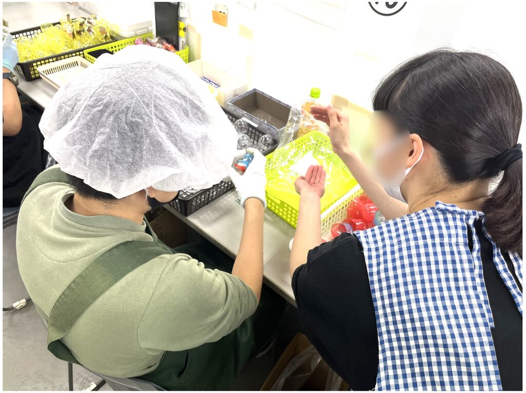 作業台の前で、ヘアキャップやエプロン、手袋を着用した人たちが、かごに入ったカプセルを扱っている様子。