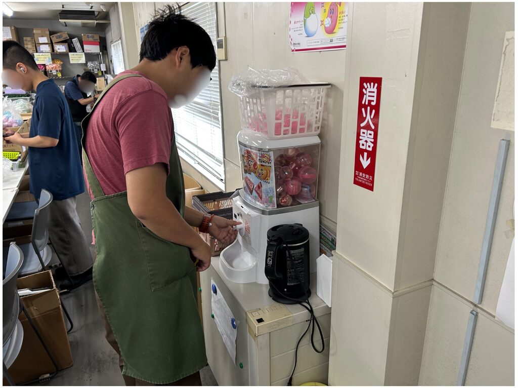 エプロンを着けた人物が、室内でカプセルトイ機のハンドルを回している。隣には黒い電気ポットが置かれ、壁には「消火器」と書かれた表示がある。