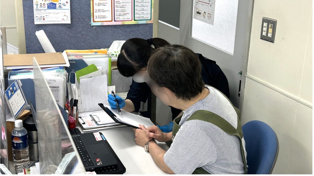 職員が来訪者に書類を見せながら説明・確認をしている様子。机上にはファイル、筆記具、書類、ノートパソコン、飲料ボトルなどが置かれている。