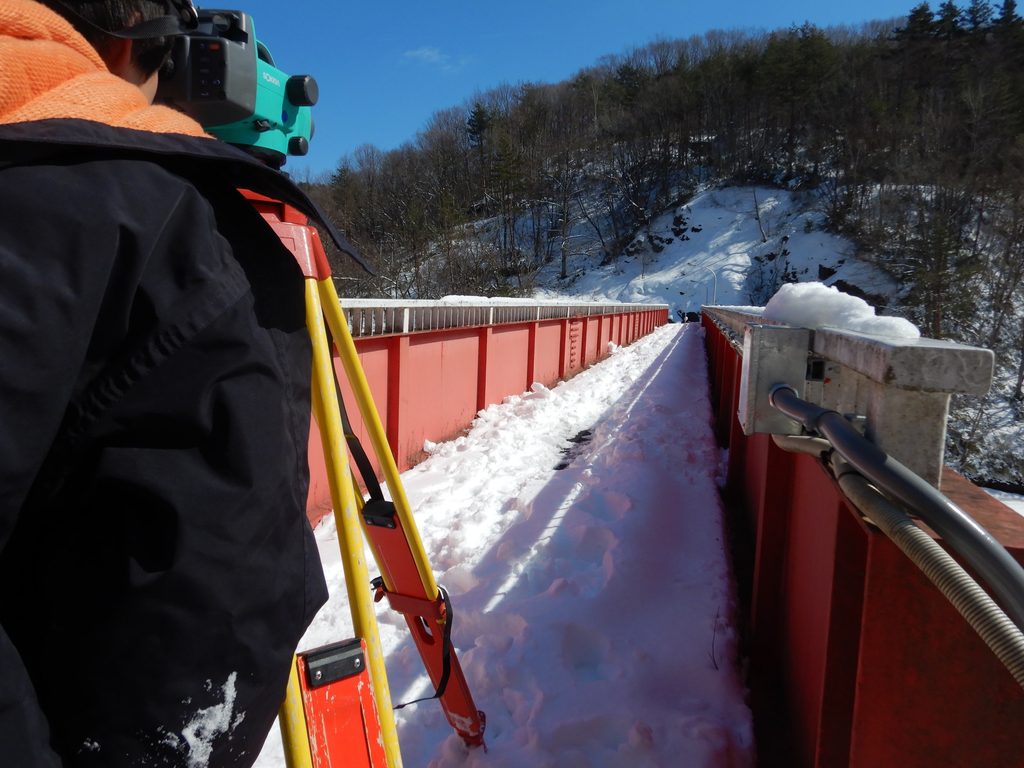 青空の下、雪の積もった赤い欄干の橋の上で、黒い上着の作業者が三脚に据えた測量機器をのぞき込んでいる。橋の路面には雪が積もり、奥に雪の斜面と林が見える。