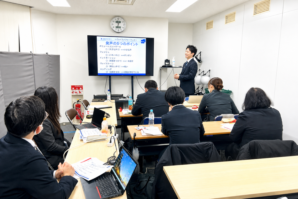 会議室で、スーツ姿の受講者が「発声の5つのポイント」を映したモニターを見ながら講師の話を聞いている研修の様子