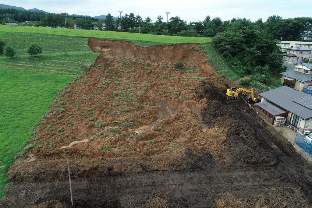 草地の斜面が広い範囲で崩れ、茶色い土がむき出しになっている。右側に黄色い重機と住宅が見える。