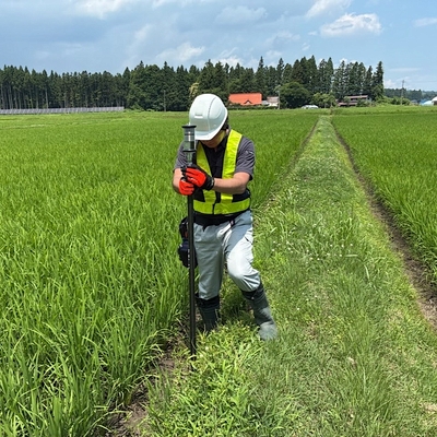 青空の下、緑の稲が育つ田んぼの畦道で、白いヘルメットと黄色のベストを着けた作業者が、縦長の棒状の機器を地面に立てて持っている。