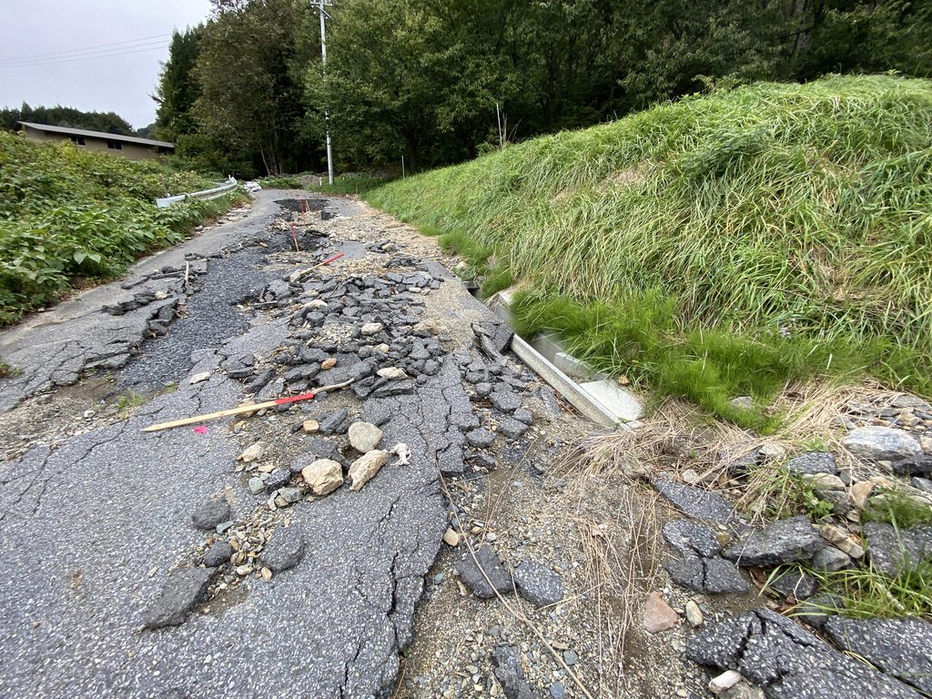 舗装がひび割れて崩れ、砕けた舗装片や石が散らばる道路。右側に草の生えた斜面と側溝があり、奥に木々と建物が見える