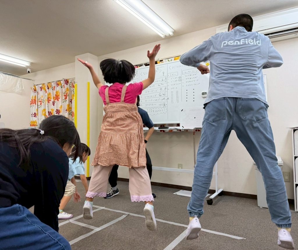 床に白いテープが貼られた室内で、両腕を上げてジャンプする子どもと、その右側で同じようにジャンプする大人の後ろ姿。左側では、数人の子どもが前かがみの姿勢をとっている。