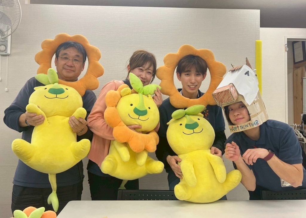 黄色い芽のキャラクターのぬいぐるみを抱え、花の形の枠から顔をのぞかせる男女4人。右端の1人は「HAPPY NEW YEAR」と書かれた段ボール製の被り物をつけている。