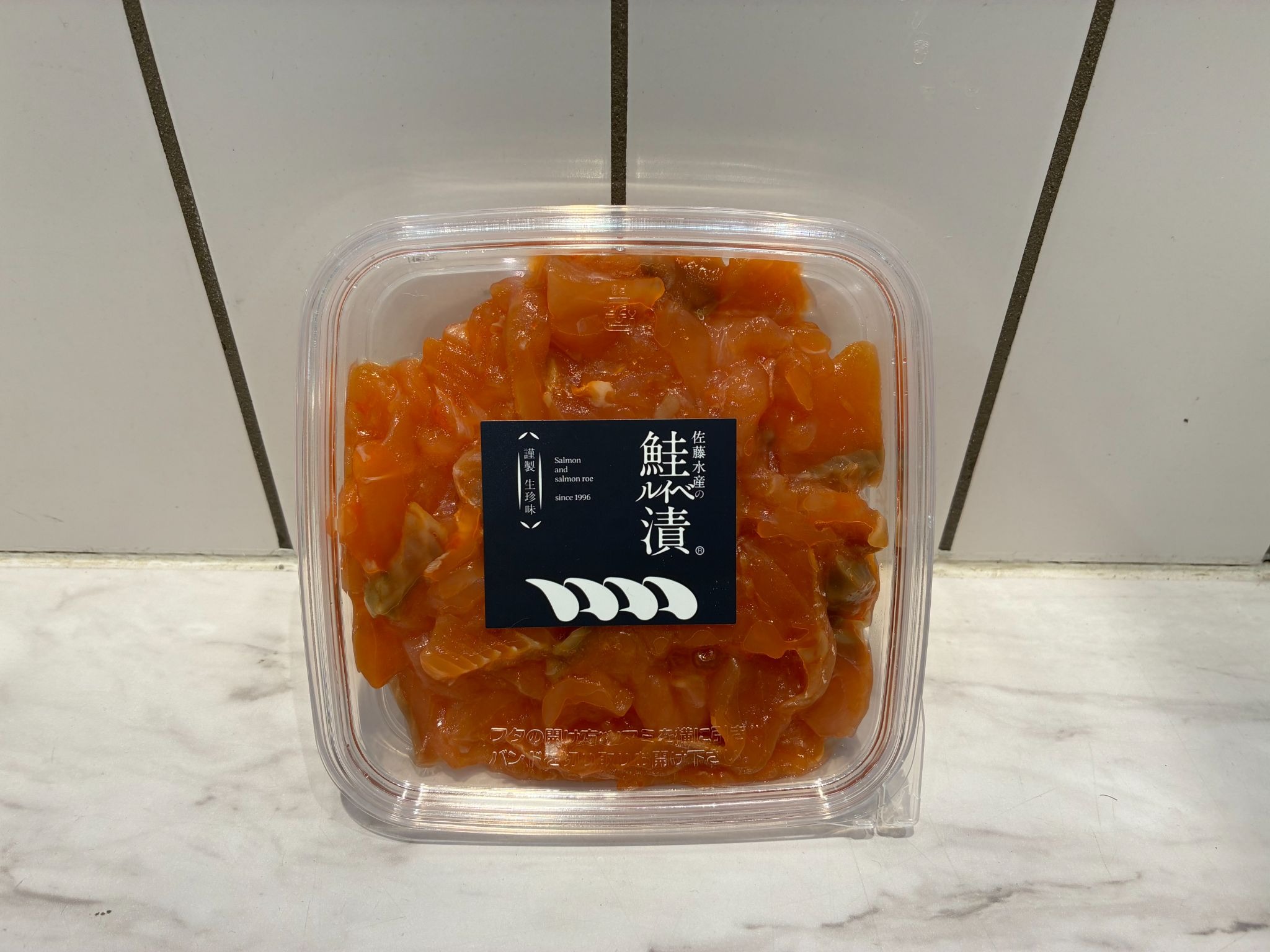 「佐藤水産の 鮭ルイベ漬」のパック。透明な容器にたれ漬けの鮭の切り身が入り、紺色のラベルが貼られている