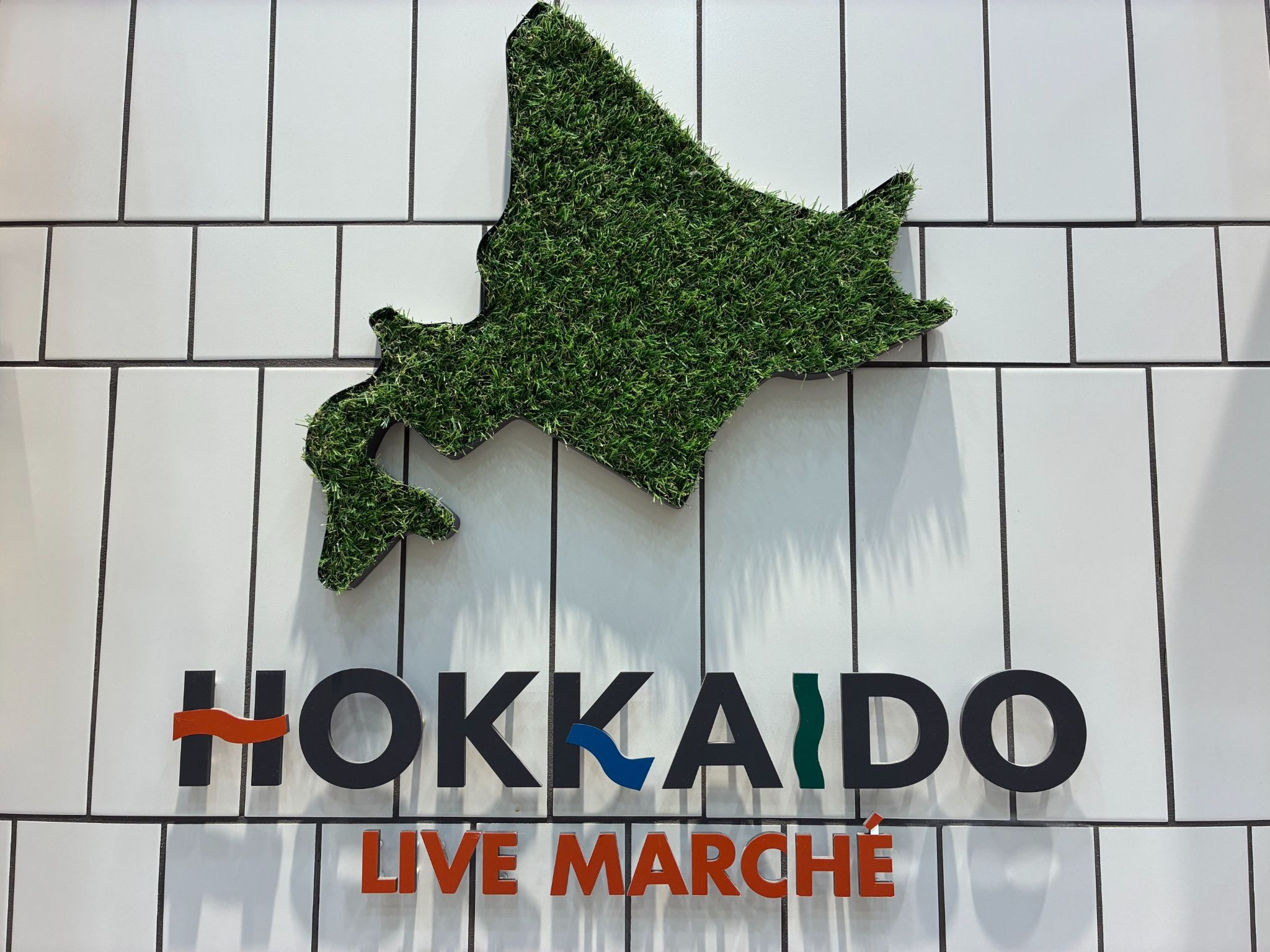 白いタイル壁に取り付けられた「HOKKAIDO LIVE MARCHÉ」の看板。人工芝で北海道の形を表したオブジェがある